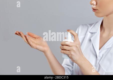 femme médecin en uniforme blanc vaporise un désinfectant sur sa main sur fond gris, gros plan. espace de copie Banque D'Images