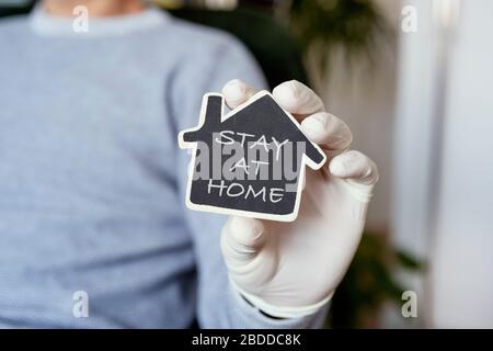 gros plan d'un homme, dans une tenue décontractée portant des gants en latex à l'intérieur, montrant le message rester à la maison écrit dans un panneau noir en forme de maison, comme mesure à Banque D'Images