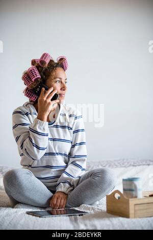 Femme avec des cheveux curlersÂ assis sur le lit et parlant sur le téléphone mobile Banque D'Images