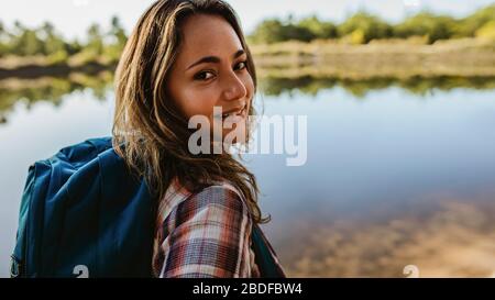Gros plan d'une belle femme avec sac à dos debout près du lac. Femme camping-car regardant l'appareil photo. Banque D'Images
