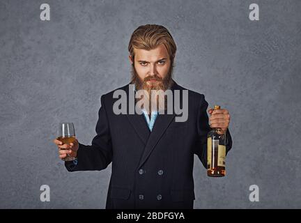 Homme barbu avec verre et bouteille de whisky. Affaires portant une chemise bleue et une veste noire. Contre la paroi grise. Banque D'Images