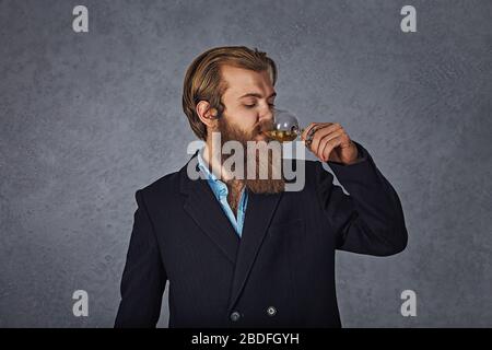 Bel homme bien habillé en barbu dans une veste tenant un verre de boisson alcool whisky isolé fond gris mur. Boissons, détente, loisirs et gens Banque D'Images