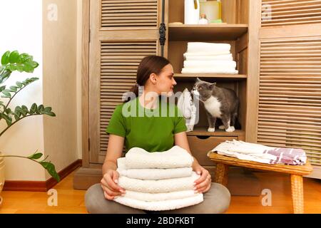 Femme portant un t-shirt vert près d'une armoire tenant des serviettes dans la buanderie avec un chat magnifique. Concept de maison confortable et propre Banque D'Images