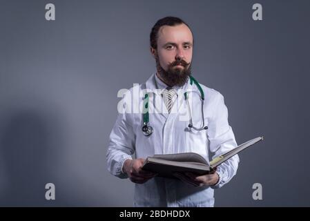 Portrait d'un médecin barbu dans un manteau blanc et un phonendoscope tenant des livres éducatifs dans ses mains. Photo Studio sur fond gris. Auto-éduca Banque D'Images