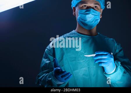 portrait de gros plan d'un chirurgien de médecin caucasien, dans un costume stérile, masque, tenant un scalpel dans ses mains, se préparant à fonctionner. Sur un fond bleu. Banque D'Images