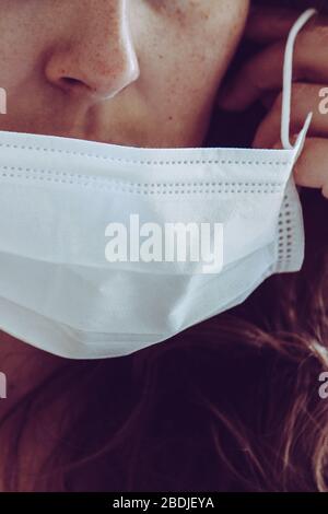 Détail du visage de femme non reconnaissable portant un masque médical blanc. Partiellement flou, mise au point sélective, grain léger. Fille REDEHEAD avec des freckles. Coronavirus, COVID-19 quarantaine. Concept médecin/infirmière. Banque D'Images