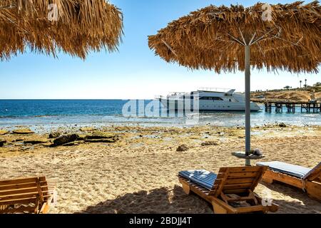 Chaises longues en bois sous un parasol en paille rugueuse sur la plage de la mer et un grand bateau à bateaux blanc dans l'eau près de la rive le jour ensoleillé de l'été. Banque D'Images