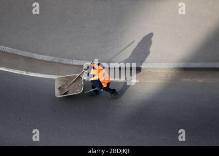 Le travailleur porte une brouette avec des ordures sur la route de la ville vide, vue du haut. Nettoyage de rue en quarantaine pendant le coronavirus covid-19 Banque D'Images