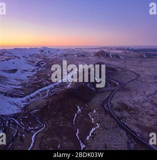 Vue sur la chaussée d'asphalte sinueuse qui traverse un terrain rocheux avec des montagnes enneigées contre le ciel coucher de soleil coloré en Islande Banque D'Images