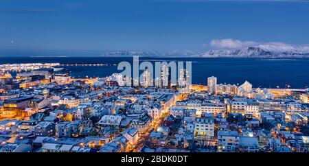 Vue aérienne de la ville éclairée sur la côte de l'océan avec des montagnes enneigées en arrière-plan à Reykjavik, en Islande Banque D'Images