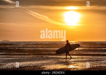 Un homme avec une planche de surf marche le long de la plage de Polzeath sur la côte nord de Cornwall pendant le verrouillage de Coronavirus. Banque D'Images