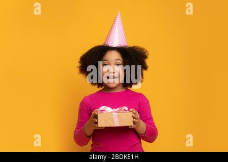 Joyeuse petite fille d'anniversaire Afro portant un chapeau de fête et un cadeau de maintien Banque D'Images