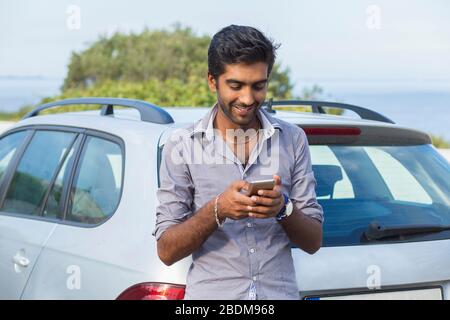 Un jeune homme indien souriant regardant le téléphone mobile. Modèle asiatique joyeux à l'extérieur à côté de sa voiture qui appelle texting. Personne Banque D'Images