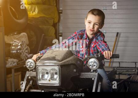 Un jeune homme jeune mécaniste automobile rêve avec joie qu'il roule rapidement sur une moto dans le garage d'une station de service. Un enfant est assis sur un vieux VTT Banque D'Images