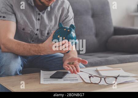 L'homme assis à une table pleine de factures impayées secoue le dernier sou de la banque de porc. Dépensez vos dernières économies. Chômage, pauvreté, faillite Banque D'Images