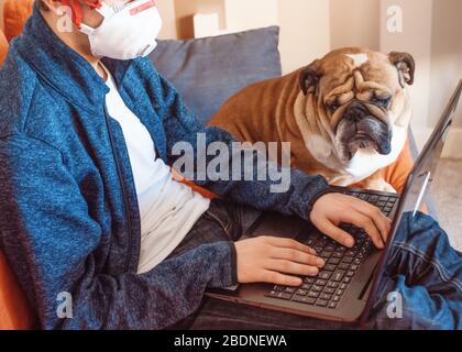 Un garçon assis sur un canapé orange portant une chemise blanche, et un Jean étudiant/apprenant en ligne sur la tablette avec son chien blanc et noir / buldog Banque D'Images