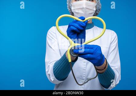 Belle femme médecin en vêtements médicaux blancs, masque tient un phonendoscope et montre un coeur, image isolée sur fond bleu Banque D'Images
