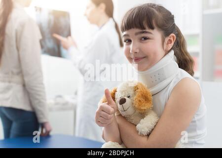 Jolie fille avec col cervical dans le bureau du médecin: Elle sourie, tenant son ours en peluche et donnant un pouce vers le haut Banque D'Images