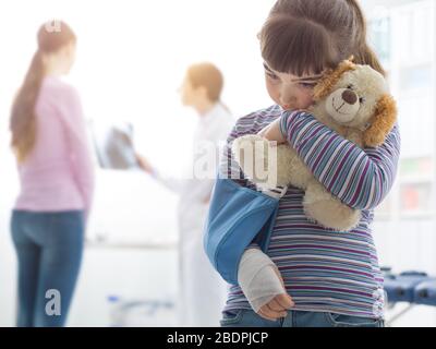 Une jolie fille effrayée avec un bras dans le cabinet du médecin, elle tient un concept d'ours en peluche Plushie, enfants et soins de santé Banque D'Images