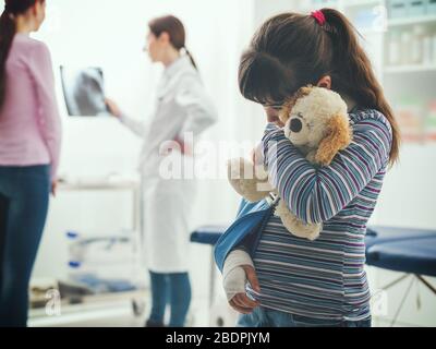 Une jolie fille effrayée avec un bras dans le cabinet du médecin, elle tient un concept d'ours en peluche Plushie, enfants et soins de santé Banque D'Images