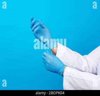 femme médecin dans un manteau blanc met sur ses mains des gants médicaux stériles en latex bleu sur ses mains, fond bleu, gros plan Banque D'Images