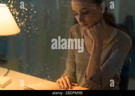 Jeune femme lisant un livre la nuit, elle est assise au bureau, verre avec des gouttes au premier plan Banque D'Images