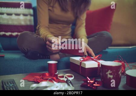 Femme préparant des cadeaux de Noël à la maison: Elle est assise sur le canapé et ajoute une carte de Noël à un présent Banque D'Images