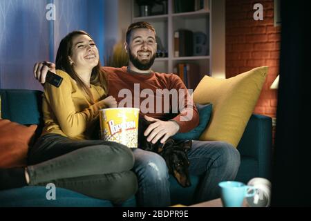 Heureux couple se reposant sur le canapé à la maison avec leur chien: Ils regardent des émissions de télévision, manger du pop-corn et rire ensemble Banque D'Images