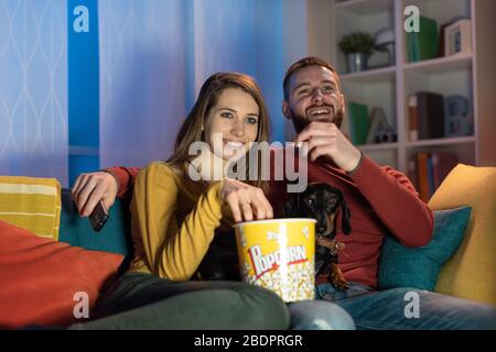 Heureux couple se reposant sur le canapé à la maison avec leur chien, ils regardent la télévision et mangent du pop-corn, il tient la télécommande Banque D'Images