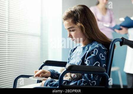 Jolie fille en fauteuil roulant à l'hôpital en utilisant un comprimé, sa mère et le médecin parlent en arrière-plan Banque D'Images