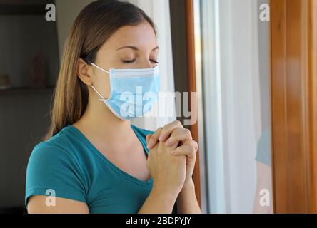 COVID-19 pandémie de coronavirus Femme priant le masque de port contre le virus SRAS-COV-2. Fille avec yeux fermés priant près de la fenêtre portant le masque chirurgical Banque D'Images