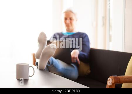 Femme assise dans un canapé avec un ordinateur portable et ses pieds sur la table où une tasse ou une tasse avec le texte ACCUEIL SWEET HOME est placé. De travail Banque D'Images