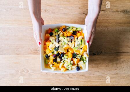 Les mains d'une femme attrapent une salade de fruits dans un bol blanc sur une table en bois Banque D'Images