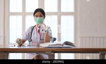 Photo de la jeune femme qui travaille comme médecin portant un stéthoscope et assis à la table en bois avec livre et tablette d'ordinateur sur l'offique ordonné du médecin Banque D'Images