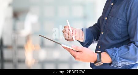 Photo d'un homme d'affaires intelligent tenant une tablette d'ordinateur et un stylet tout en se tenant dans le bureau confortable avec un mur de verre flou de bureau comme backgro Banque D'Images