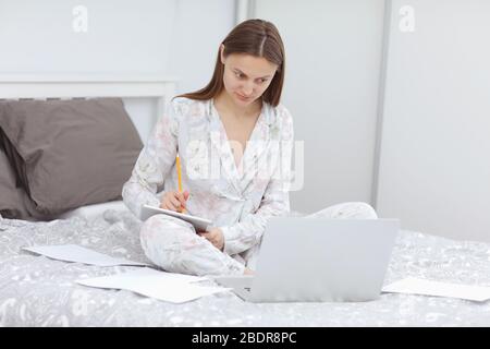 Une belle femme d'affaires qui travaillait sur un ordinateur portable sur le lit. Une fille qui s'est mise en quarantaine au bureau à domicile. Restez à la maison. Freelance. Communication et Banque D'Images