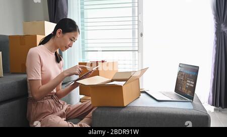 Photo de la jeune belle femme emballant ses marchandises pour livraison au client en utilisant une tablette d'ordinateur et un ordinateur portable dans le salon confortable. Démarrer Banque D'Images