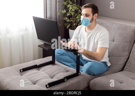 Jeune homme caucasien travaillant à domicile portant un masque de protection à l'aide d'un ordinateur portable et d'Internet. Confortable bureau à la maison, lieu de travail sur canapé pendant le coronavirus Banque D'Images