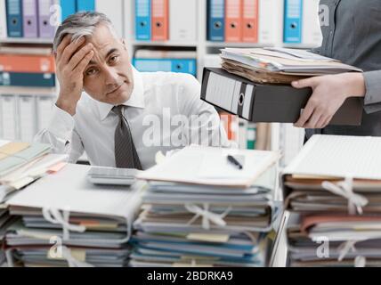 Souligné businessman working at office desk et débordé de travail, le bureau est couvert de paperasse, sa secrétaire est ce qui porte plus de fichiers Banque D'Images