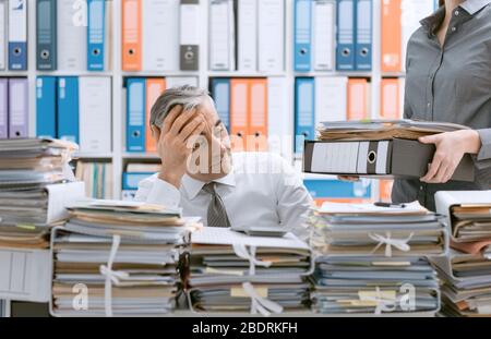 Souligné businessman working at office desk et débordé de travail, le bureau est couvert de paperasse, sa secrétaire est ce qui porte plus de fichiers Banque D'Images