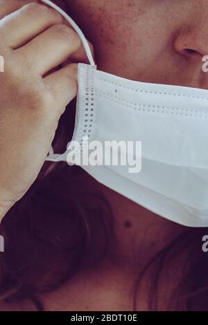 Détail du visage de femme non reconnaissable portant un masque médical blanc. Partiellement flou, mise au point sélective, grain léger. Fille REDEHEAD avec des taches. Coronavirus, COVID-19 quarantaine. Médecins, concept infirmière. Banque D'Images