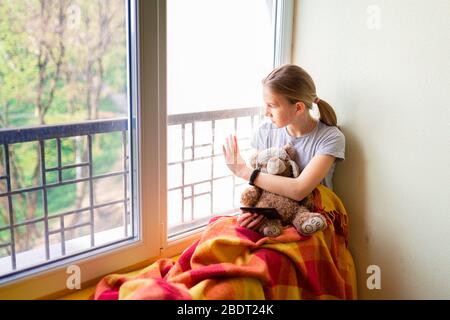 Une fille triste assise sur la fenêtre avec un jouet à la maison isolée, en regardant dehors. Concept de quarantaine du coronavirus. Banque D'Images