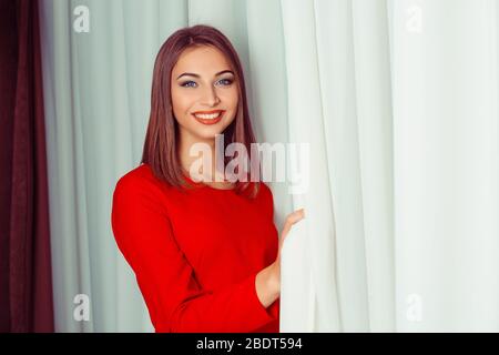 Portrait d'une jeune femme souriante dans des rideaux d'ouverture de robe rouge. Prise de vue en intérieur, image horizontale Banque D'Images