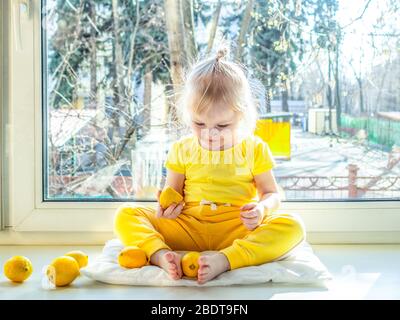 Une petite fille en vêtements jaunes se trouve sur un seuil de fenêtre pendant la journée et sourit. Fille tenant les jambes de citron. Un soleil brillant brille dans le dos de la fille. Banque D'Images