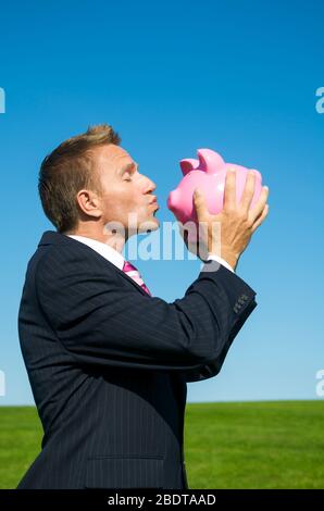 Happy businessman kissing pink piggy bank à l'extérieur sur pré vert lumineux avec ciel bleu copy space Banque D'Images