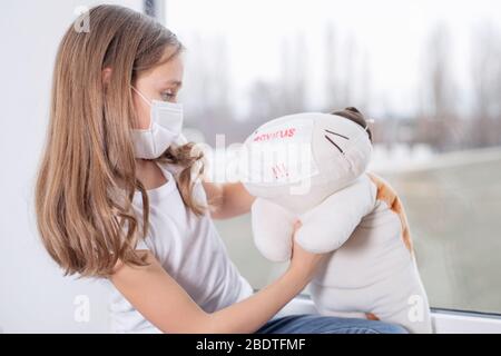 L'épidémie de coronavirus dans le monde entier.UNE fille triste dans un masque protecteur contre les virus sur son visage est assise par la fenêtre et joue avec un jouet. Banque D'Images