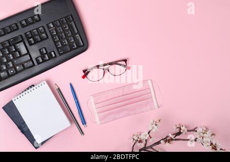 Branches fleuries avec un masque médical, un clavier, un bloc-notes et des lunettes sur un fond rose. Coronavirus COVID-19 Banque D'Images