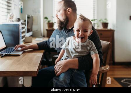 Le père essaie de travailler de chez lui avec un enfant hurlant sur ses genoux Banque D'Images