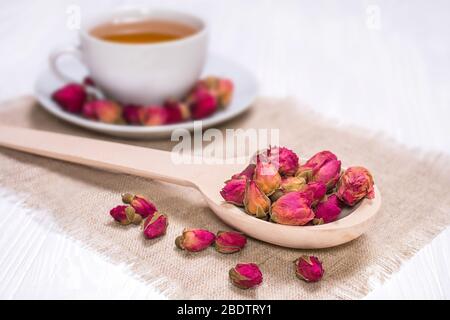 Thé de fines herbes avec rose. Tasse blanche avec du thé chaud. Une cuillère en bois avec des boutons de fleurs séchées sur une serviette en lin. Banque D'Images