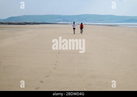 Les résidents de Borth, la mère à vélo et le fils en course, font leur exercice quotidien pendant le verrouillage de Coronavirus Covid 19. Sur la plage de Ynyslas /Borth à marée basse.cette station balnéaire était vide à part quelques habitants.la police a encouragé/imposé aux non-locaux de rester loin. Banque D'Images